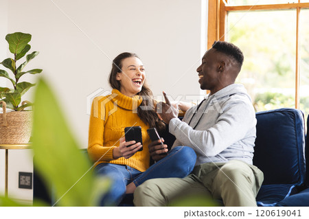 Happy diverse couple sitting on sofa using smartphones at home 120619041