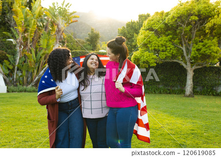 Three happy diverse female friends with flag on backs in sunny garden Three happy diverse female friends with flag on backs in sunny garden 120619085