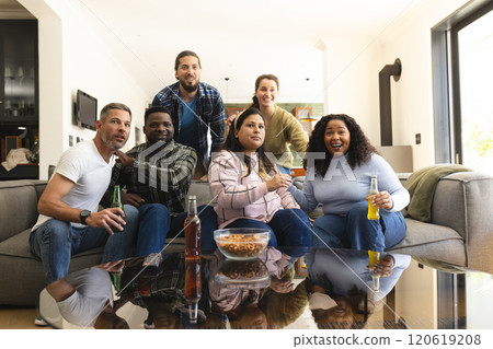 Happy diverse male and female friends watching sport on tv at home and drinking beers, copy space 120619208