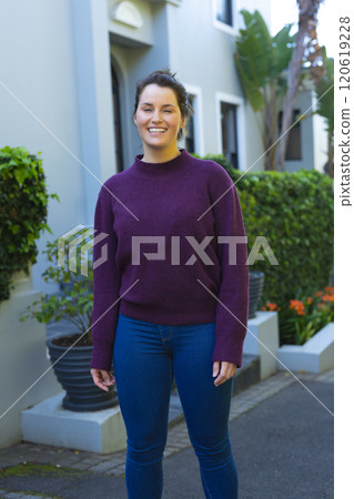 Happy caucasian woman in purple sweater standing and smiling in front of house in garden 120619228