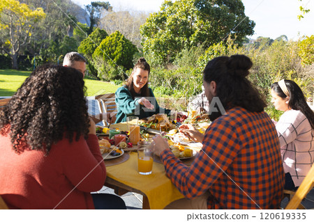 Happy diverse male and female friends eating thanksgiving celebration meal in sunny garden 120619335