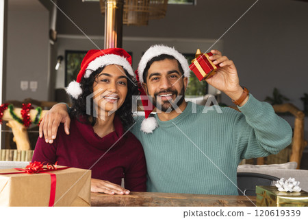 Happy biracial couple with santa hats embracing and smiling among gifts at christmas at home 120619339