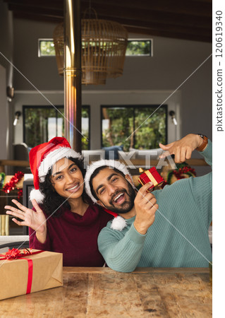 Happy biracial couple with santa hats embracing and laughing among gifts at christmas at home 120619340