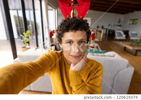 Portrait of happy biracial man wearing reindeer antlers and having video at christmas at home 120619374