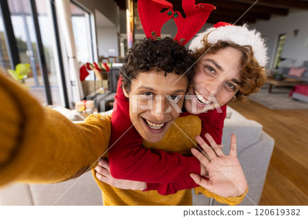 Portrait of happy diverse gay male couple wearing christmas hats and having video call at home 120619382