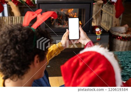 Diverse gay male couple wearing christmas hats and having smartphone video call at home, copy space Diverse gay male couple wearing christmas hats and having smartphone video call at home, copy space 120619384