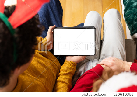 Diverse gay male couple wearing christmas hats and having tablet video call at home, copy space Diverse gay male couple wearing christmas hats and having tablet video call at home, copy space 120619395