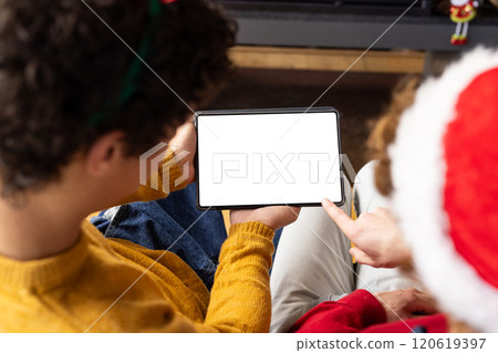 Diverse gay male couple wearing christmas hats and having tablet video call at home, copy space Diverse gay male couple wearing christmas hats and having tablet video call at home, copy space 120619397