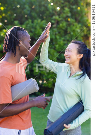 Happy diverse couple holding yoga mats high fiving in sunny garden 120619411