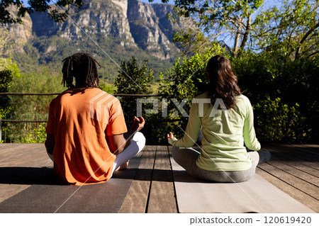 Rear view of diverse couple practicing yoga meditation sitting on deck in sunny garden 120619420