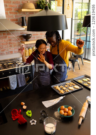 Happy diverse couple in aprons baking christmas cookies, dancing in kitchen Happy diverse couple in aprons baking christmas cookies, dancing in kitchen 120619486