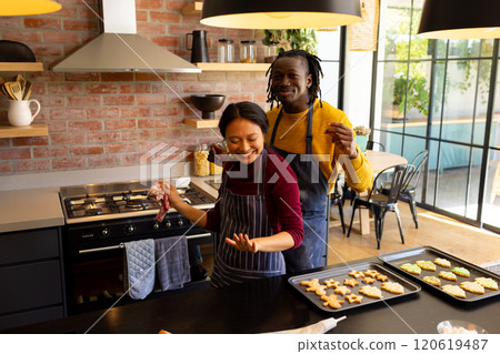 Happy diverse couple in aprons baking christmas cookies, dancing in kitchen, copy space 120619487