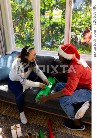 Happy diverse couple sitting on couch wrapping christmas gifts in sunny living room, copy space Happy diverse couple sitting on couch wrapping christmas gifts in sunny living room, copy space 120619496