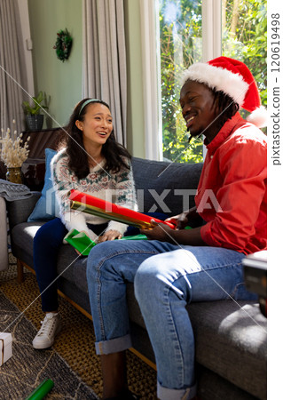 Happy diverse couple sitting on couch wrapping christmas gifts in sunny living room 120619498