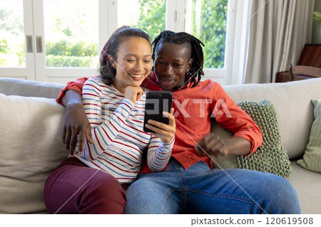 Happy diverse couple using smartphone together sitting on couch smiling in sunny living room Happy diverse couple using smartphone together sitting on couch smiling in sunny living room 120619508