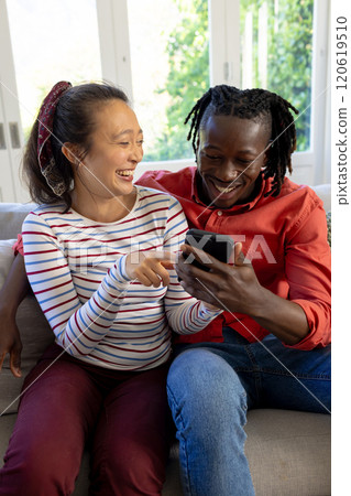 Happy diverse couple using smartphone together, smiling in sunny living room 120619510