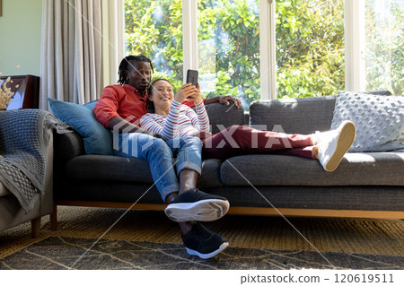 Happy diverse couple using smartphone together relaxing on couch in sunny living room Happy diverse couple using smartphone together relaxing on couch in sunny living room 120619511