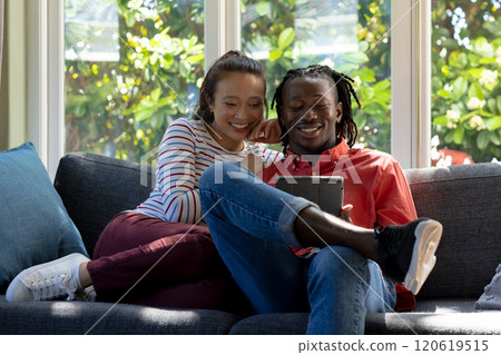 Happy diverse couple using tablet together sitting on couch smiling in sunny living room Happy diverse couple using tablet together sitting on couch smiling in sunny living room 120619515