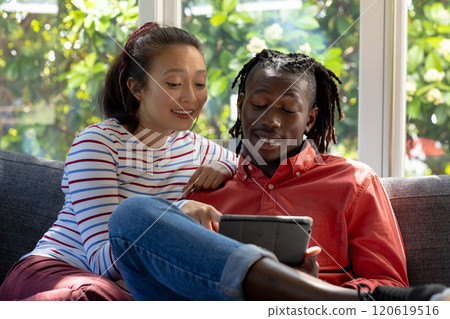 Happy diverse couple using tablet together sitting on couch smiling in sunny living room Happy diverse couple using tablet together sitting on couch smiling in sunny living room 120619516