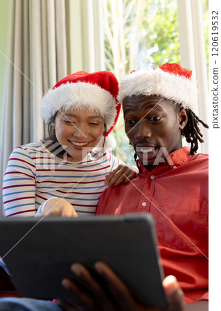 Happy, surprised diverse couple in christmas hats having tablet video call in sunny living room Happy, surprised diverse couple in christmas hats having tablet video call in sunny living room 120619532