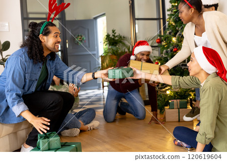 Wearing festive hats, multiracial friends exchanging gifts by christmas tree, at home Wearing festive hats, multiracial friends exchanging gifts by christmas tree, at home 120619604
