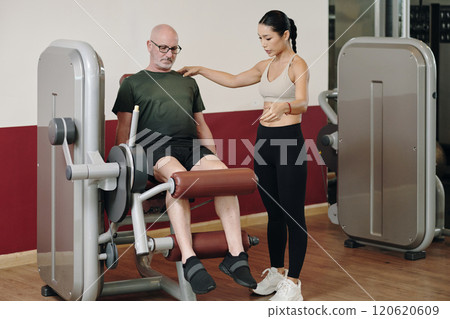 Helping Senior Man Exercising on Gym Equipment with Trainer 120620609
