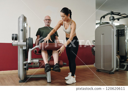 Assisting Senior Man with Leg Exercise Machine in Gym 120620611