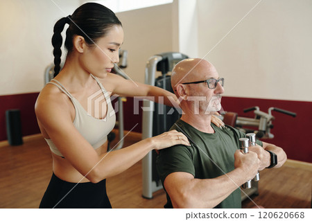 Assisting Senior Man Lifting Weights in Gym Setting 120620668