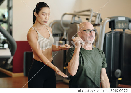 Personal Trainer Assisting Senior Client in Gym 120620672