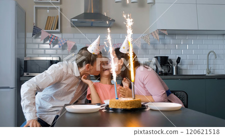 Father and mother in festive hats kiss their daughter while admiring the sparkling ice fountains on a birthday cake Father and mother in festive hats kiss their daughter while admiring the sparkling ice fountains on a birthday cake 120621518