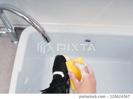 Hand-cleaning black sneakers with a yellow sponge under running water in a sink, demonstrating effective footwear care. 120624367