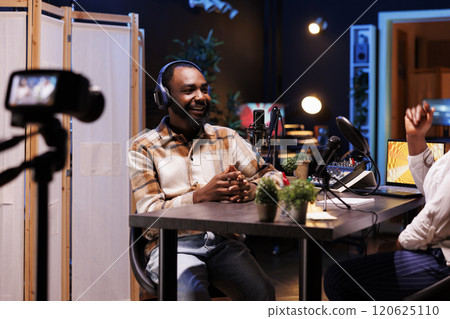 African American man creates vlogs using digital camera while producing podcasts with professional audio gadgets. Male journalist interviews guest in his home studio, filming with device on a tripod. 120625110