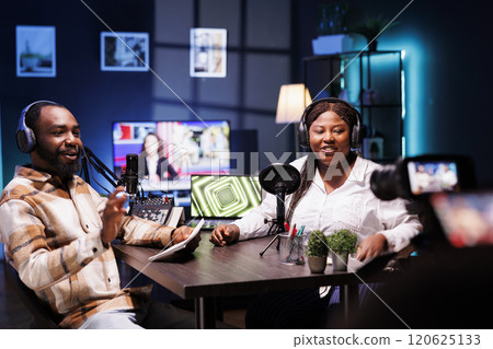 African american host using device on tripod to record entertaining discussion with woman during livestream in apartment studio. Presenter uses professional camera to produce comedy podcast. African american host using device on tripod to record entertaining discussion with woman during livestream in apartment studio. Presenter uses professional camera to produce comedy podcast. 120625133