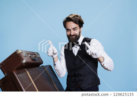 Young bellboy dancing on camera and showing groovy moves against blue background, acting cheerful and funny. Stylish trendy doorkeeper fooling around next to trolley bags, having fun. Young bellboy dancing on camera and showing groovy moves against blue background, acting cheerful and funny. Stylish trendy doorkeeper fooling around next to trolley bags, having fun. 120625135