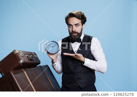 Young doorman poses with wall clock, looking at time and panicking for being late on hospitality industry occupation. Punctual hotel porter using watch to see exact hour and minutes. Young doorman poses with wall clock, looking at time and panicking for being late on hospitality industry occupation. Punctual hotel porter using watch to see exact hour and minutes. 120625278