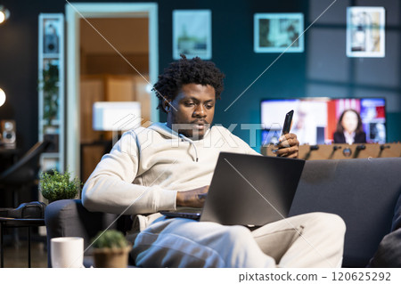 Man sitting on couch in cozy living room, watching internet clips on laptop. African american person enjoying leisure time in RGB illuminated apartment, looking at online videos 120625292