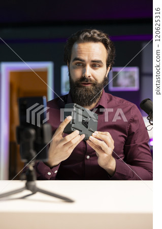 Tech expert filming video review of portable V mount battery using cellphone on tripod, feeling unconvinced of performance. Man analyzing lithium li-ion charging gadget, recording with mobile phone Tech expert filming video review of portable V mount battery using cellphone on tripod, feeling unconvinced of performance. Man analyzing lithium li-ion charging gadget, recording with mobile phone 120625316