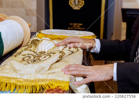 Person carefully handles beautifully adorned Torah scroll as preparations for religious service take place inside synagogue atmosphere is respectful solemn. 120627556