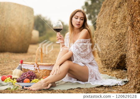 Beautiful woman with red wine in hand and wearing in white transparent dress with basket of apples in field sitting by haystack Beautiful woman with red wine in hand and wearing in white transparent dress with basket of apples in field sitting by haystack 120629128