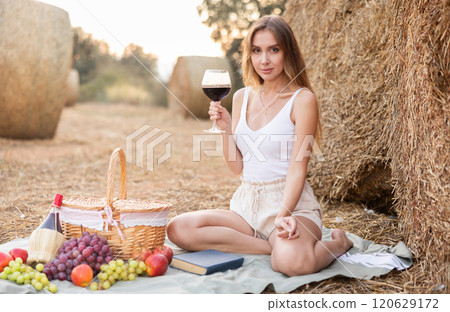 Young pretty woman in white blouse and shorts sitting on soft mat near stack of straw with glass of wine, romantic picnic in field 120629172