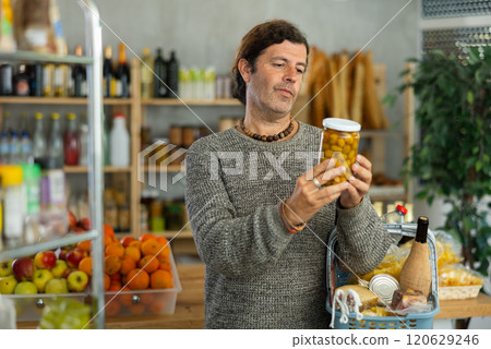 Man customer with shopping cart or basket buying food in grocery store, reading ingredients on jar of conserve olives Man customer with shopping cart or basket buying food in grocery store, reading ingredients on jar of conserve olives 120629246