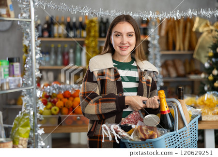 Portrait of female customer with cart choosing food and wine for Christmas celebration in grocery department of supermarket 120629251