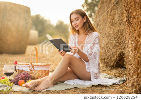 Woman in transparent dress and white lingerie reading book on picnic near hay bale 120629254