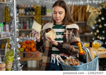 Portrait of young girl buyer with shopping cart or basket holding assortment of cheese choosing for christmas table in grocery shop Portrait of young girl buyer with shopping cart or basket holding assortment of cheese choosing for christmas table in grocery shop 120629307