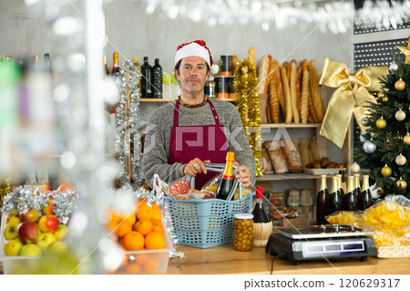 Adult male seller in grocery store posing behind counter with basket full of products for Christmas table Adult male seller in grocery store posing behind counter with basket full of products for Christmas table 120629317
