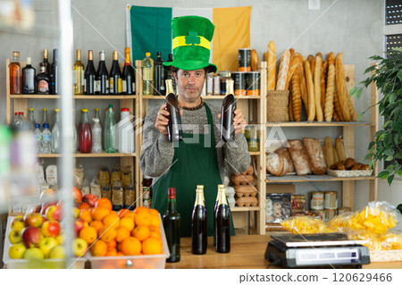 Adult man seller posing in green costume and hat, offering bottle of whiskey in store decorated for St. Patrick's Day Adult man seller posing in green costume and hat, offering bottle of whiskey in store decorated for St. Patrick's Day 120629466