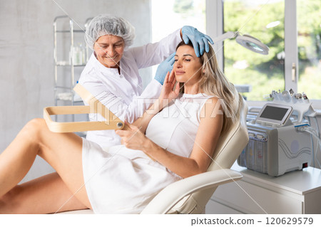 Young woman looking in mirror while mature cosmetologist showing results of facial procedure 120629579