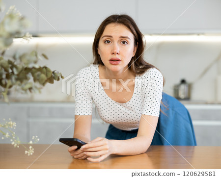 Middle-aged woman leaning on kitchen table waiting for call on smartphone Middle-aged woman leaning on kitchen table waiting for call on smartphone 120629581