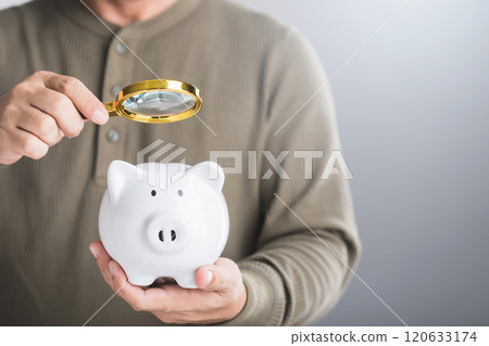 A man holds a white piggy bank and uses a magnifying glass to examine it, symbolizing the careful study of savings, financial planning, and investment strategies for future growth A man holds a white piggy bank and uses a magnifying glass to examine it, symbolizing the careful study of savings, financial planning, and investment strategies for future growth 120633174