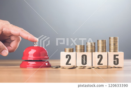 Hand presses a red bell next to a stack of coins on a wooden block labeled '2025,' representing financial goals and future planning with a sense of urgency for the upcoming year Hand presses a red bell next to a stack of coins on a wooden block labeled '2025,' representing financial goals and future planning with a sense of urgency for the upcoming year 120633390